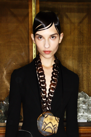 Ein Model steht vor einer braunen Schrankwand. Sie trägt einen schwarzen Blazer und eine XL-Halskette. Sie befindet sich im Backstage-Bereich bei der S/S 2017 Modenschau von Givenchy. Ihr Haare sind im burschikosen Wet-Look gestylt.