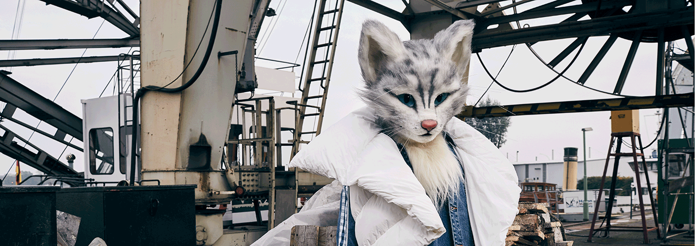 Die Katzendame Pheline trägt eine weiße Daunenweste und befindet sich im urbanen Raum am Hafen. Dazu kombiniert sie eine Jeanshose und ein Jeansoberteil.