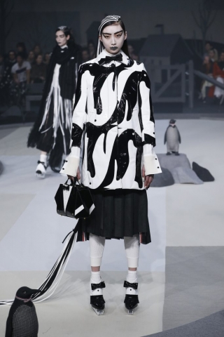 Auf dem Bild ist ein Model bei der Herbst/Wintermodenschau 2017 von Designer Thom Browne zu sehen. Sie trägt eine schwarz-weiße Jacke mit grafischem Pinguin-Print. Dazu kombiniert sie eine weiße Bluse mit überlangen Manschetten, eine schwarze Fliege,einen schwarzen Faltenrock, eine weiße Leggins und Schlittschuhe. Ihr Gesicht ist weiß geschminkt.