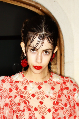 Auf dem Foto ist ein dunkelhaariges Model im Backstage-Bereich der Fashion Show von Designerin Simone Rocha zu sehen. Sie hat den Mund geschlossen und trägt roten, glossy Lippenstift.