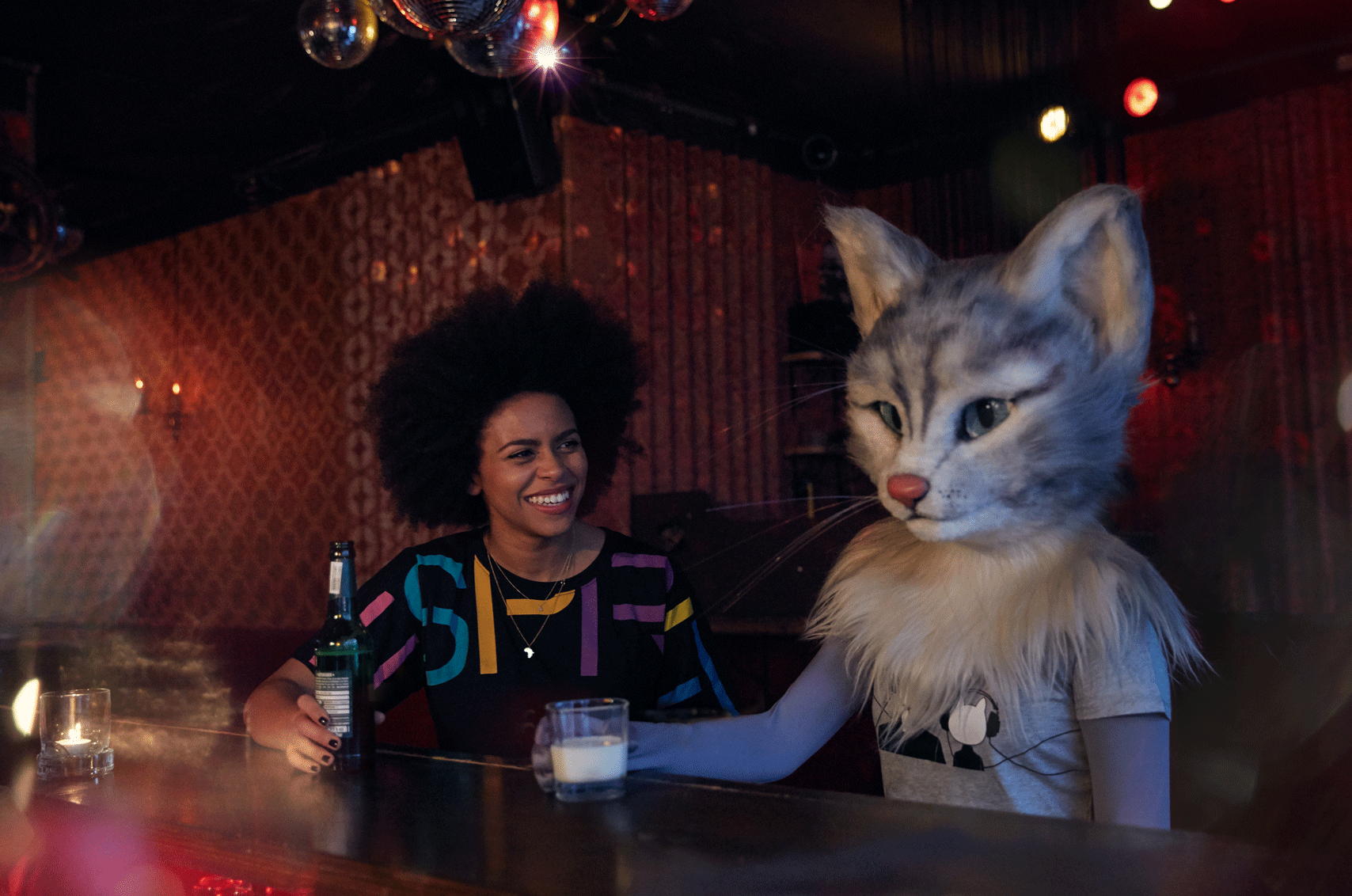 Auf dem Foto sind Aminata Belli und Katzenlady Pheline zu sehen. Die beiden sitzen an einer Bar, sprechen über Musik und eine Playlist und lauschen einem Song.