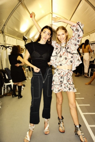 Auf dem Foto sind zwei Models im Backstagebereich der F/S-Modenschau 2017 von DEsignerin Isabel Marant zu sehen. Das linke Mädchen trägt eine schwraze 7/8 Hose mit weißen Ziernähten. Dazu kombiniert sie ein schwarzes T-Shirt uber dem sie einen schwarzen Underwear Satin-Bra trägt.