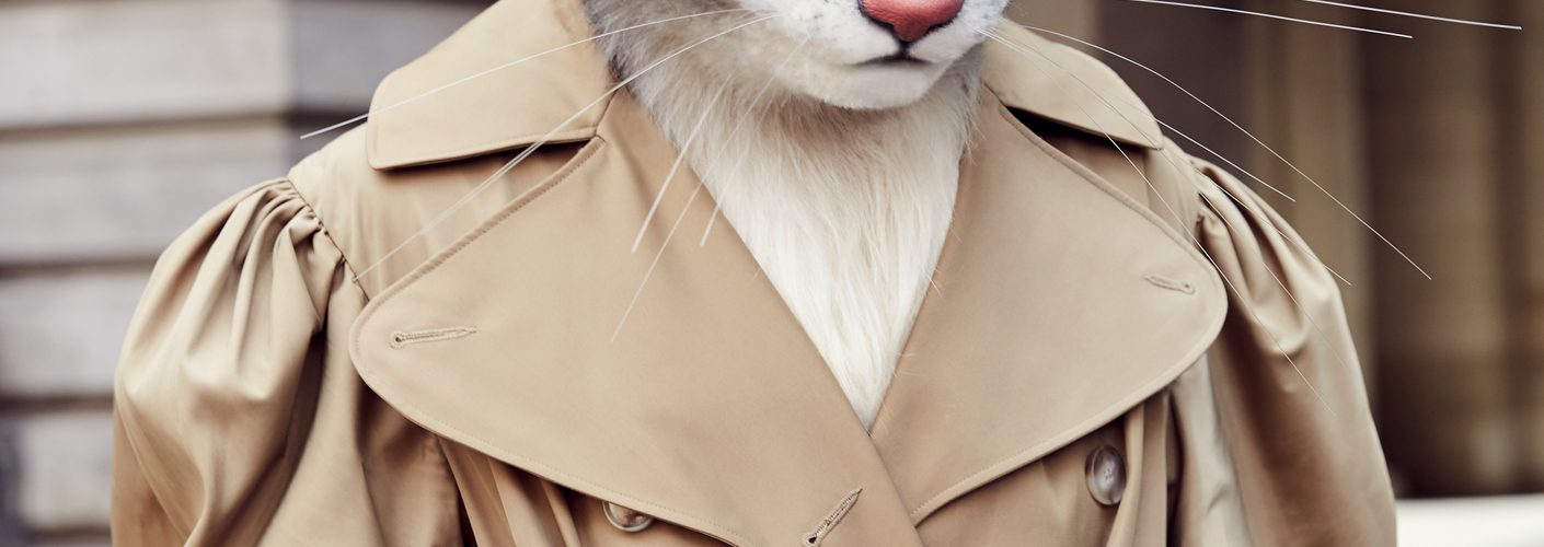 Auf dem Foto ist Katzenlady Pheline stehend vor einem sandsteinfarbenen Gebäude zu sehen. Sie trägt einen Trenchcoat mit halben Puffärmeln von Simone Rocha.