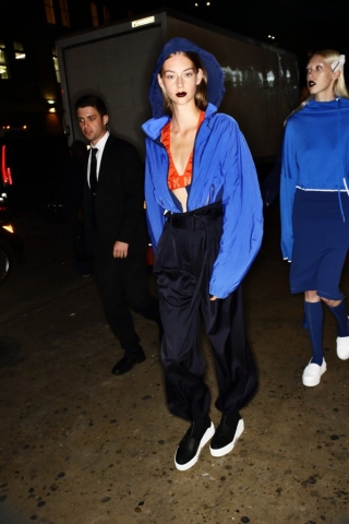 Auf dem Foto ist ein Model im Backstagebereich bei der Frühjahr/Sommermodenshow 2017 von DKNY zu sehen. Das Model trägt einen blauen Blouson mit einer Kapuze, eine shiny Jogginghose, Plateau-Sneakers und ein orangefarbenes Top mit Logo-Print.