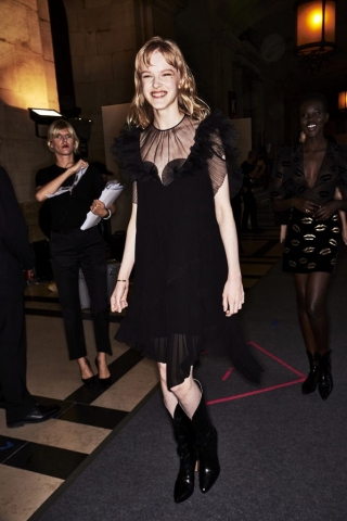 Auf dem Foto ist ein Model im Backstage-Bereich der ersten Modenschau von Designerin Claire Waight Keller für das Modehaus Givenchy in Paris zu sehen. Es handelt sich um die Präsentation der F/S 2018 Kollektion. Das Model tägt ein schwarzes Kleid mit kurzen Rüschenärmeln. Dazu kombiniert sie schwarze Cowboystiefel..