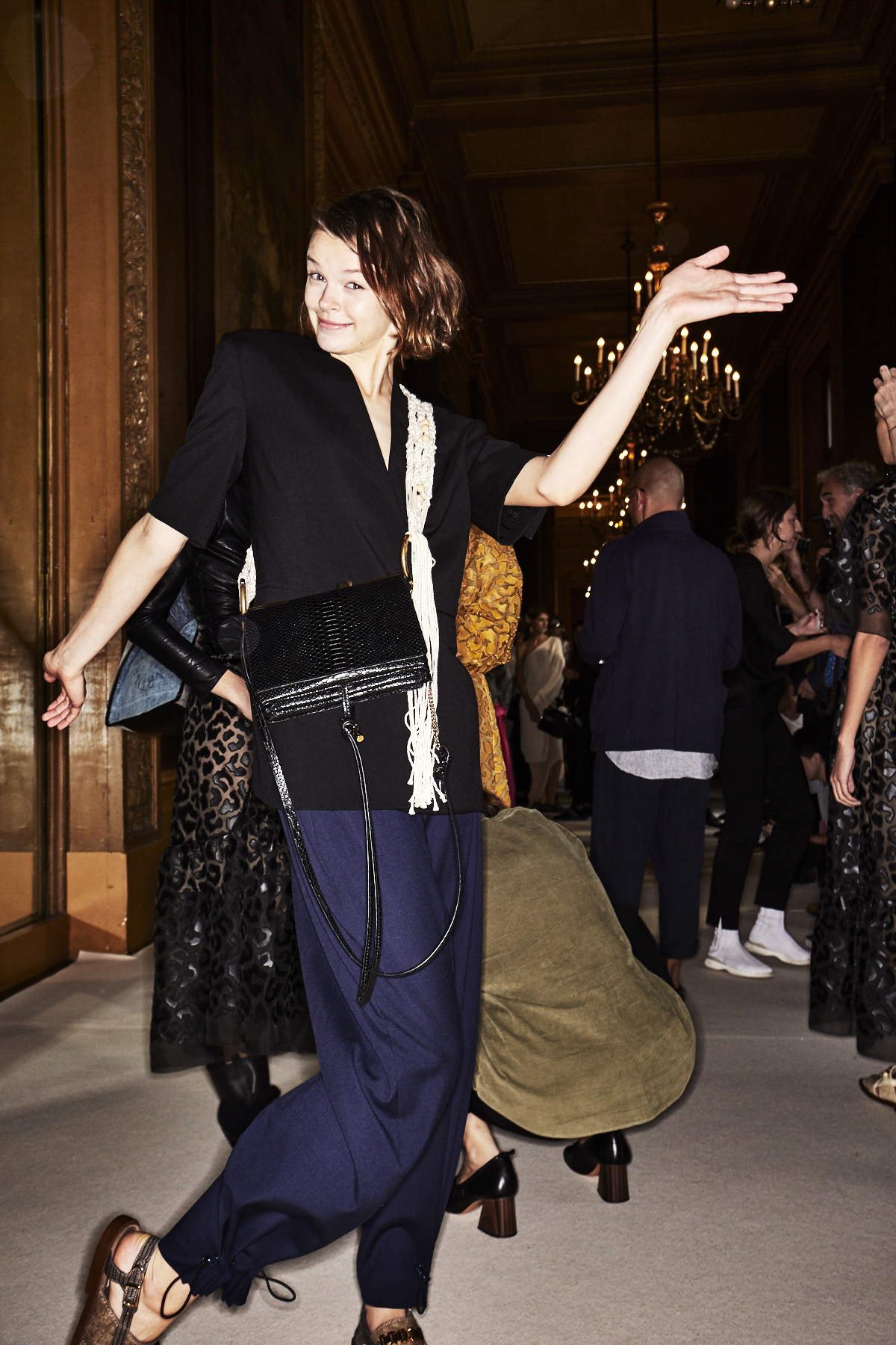 Auf dem Foto ist ein Model im Backstage Bereich bei der Frühjahr/Sommermodenschau 2018 von Designerin Stella Mc Cartney zu sehen. Sie kombiniert die Trendfarben Schwarz und Blau in Form eines halbarm Blazers und einer dunkelblauen Hose miteinander. Dazu kombiniert sie eine schwarze Crossover-Bag und einen weißen Schal.