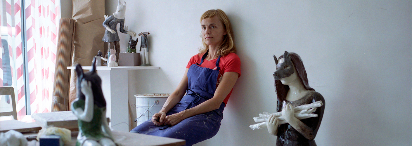 Auf dem Foto ist die französische Keramikkünstlerin Clémentine de Chabaneix in ihrem Pariser Atelier auf einem Hocker sitzend gegen die gelehnt zu sehen. Um sie herum befinden sich mehrere ihrer Keramik-Skulpturen. Das Foto stammt von Fanny Begoin.