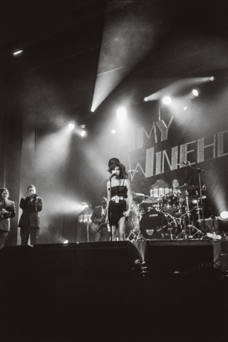 Amy Winehouse steht auf einer Bühne vor einem Mikrofon. Sie trägt ein schwarzes Minikleid. Hinter ihr ist ein riesiges Schlagzeug zu erkennen.